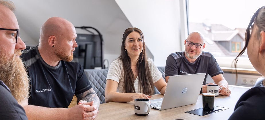 Teammeeting mit fünf Mitarbeitern von wodacon, Laptops und Tassen auf einem Tisch in hellem, modernem Raum.