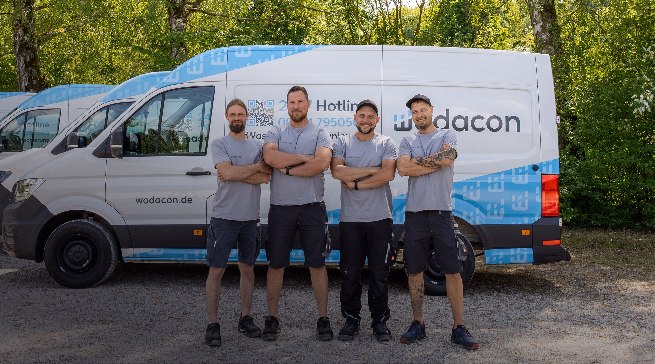 Team von wodacon vor Fahrzeugen, einheitliche graue T-Shirts, entspannte Pose, grüne Baumkulisse